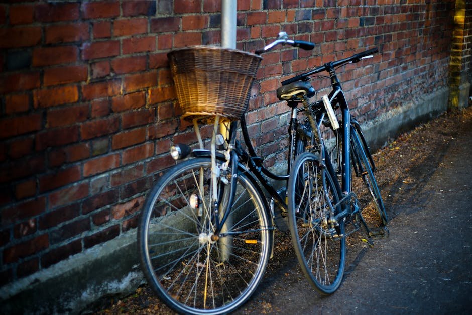 Two vintage bicycles with a basket leaning against a rustic brick wall in daylight.